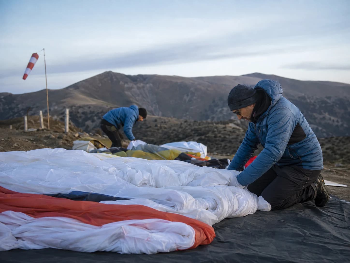 Two people prepare a paraglider beside a windsock on a mountain launch.